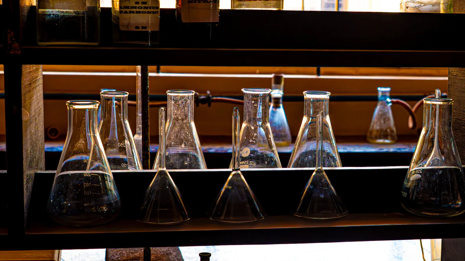 Glass conical flasks, funnels and reagent bottles on a backlit lab shelf