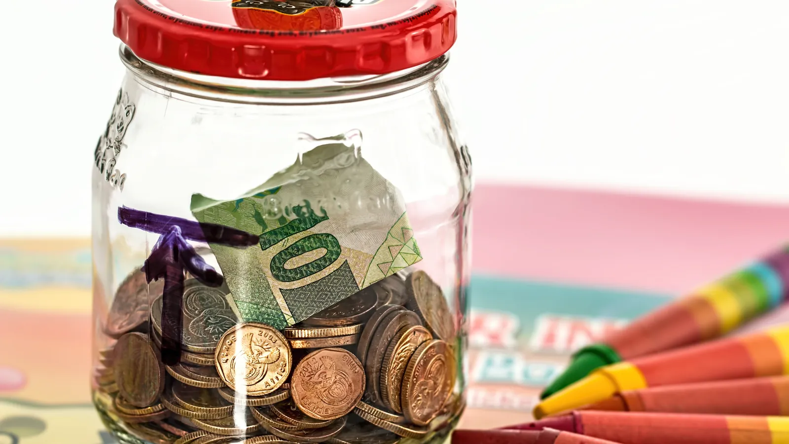 Glass jar of coins and a banknote beside colourful crayons