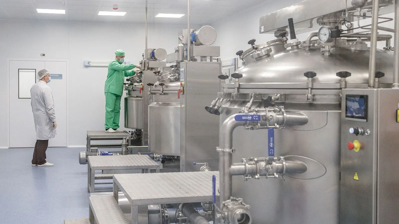 Gowned workers beside steel mixing tanks in a pharmaceutical production hall