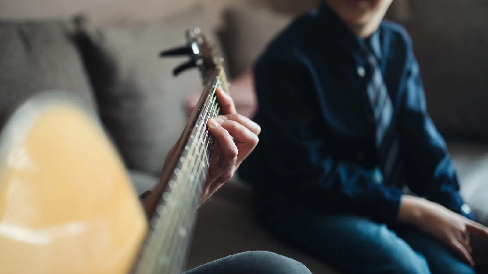 Hand fretting an acoustic guitar while a young boy on a sofa watches