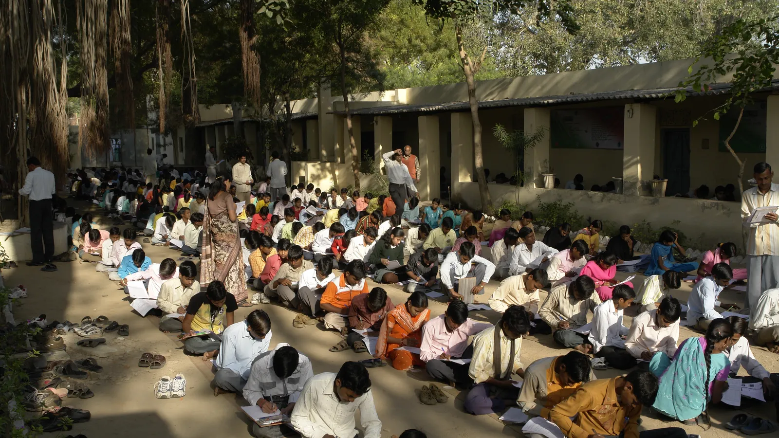Indian students sitting on the ground in a shaded school courtyard writing an exam