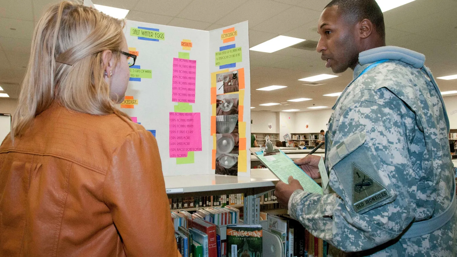 Judge in uniform with a clipboard questions a student beside a 'Salt Water Eggs' science fair board