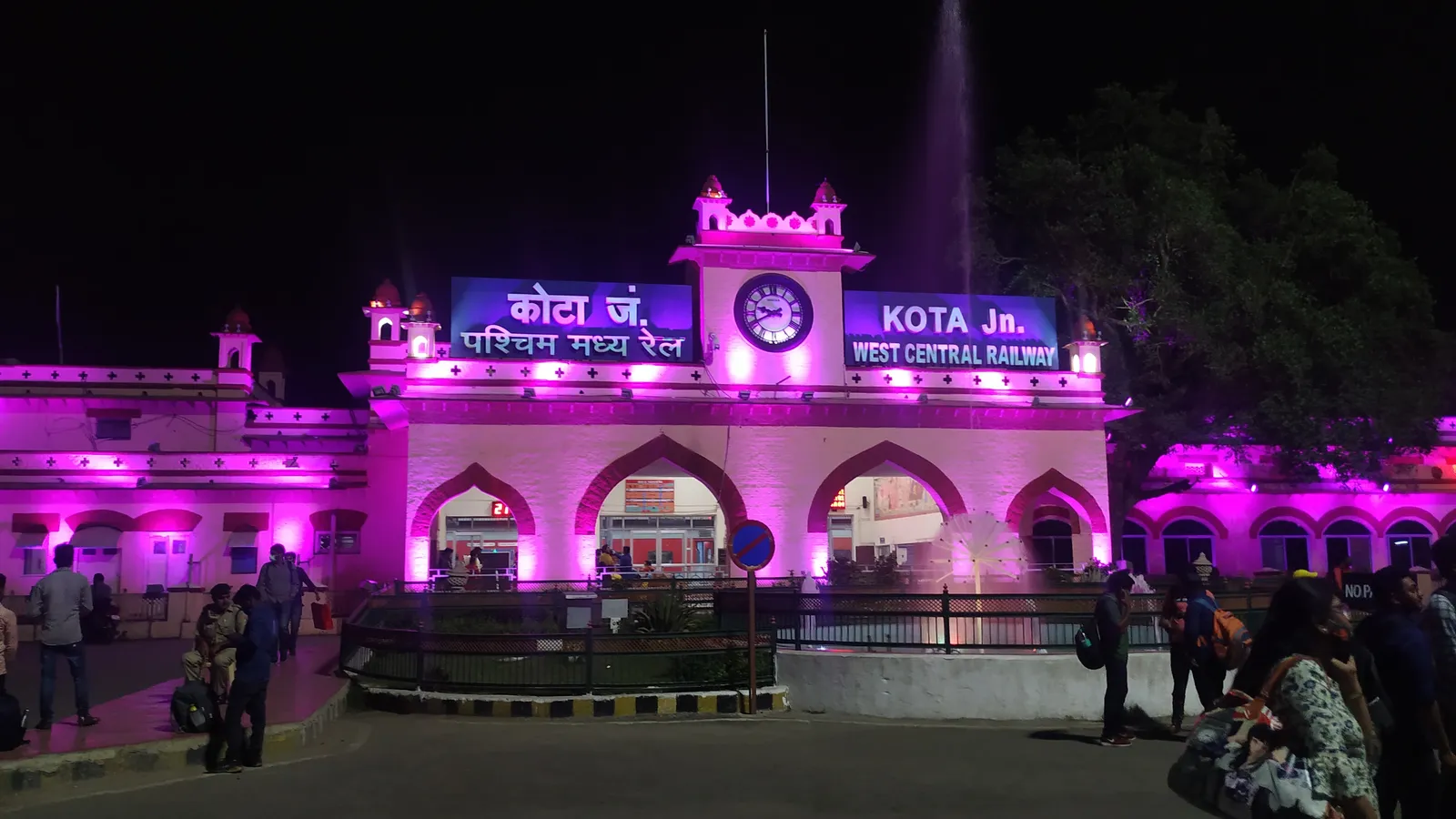 Kota Junction railway station lit up in pink at night, with travellers carrying backpacks outside