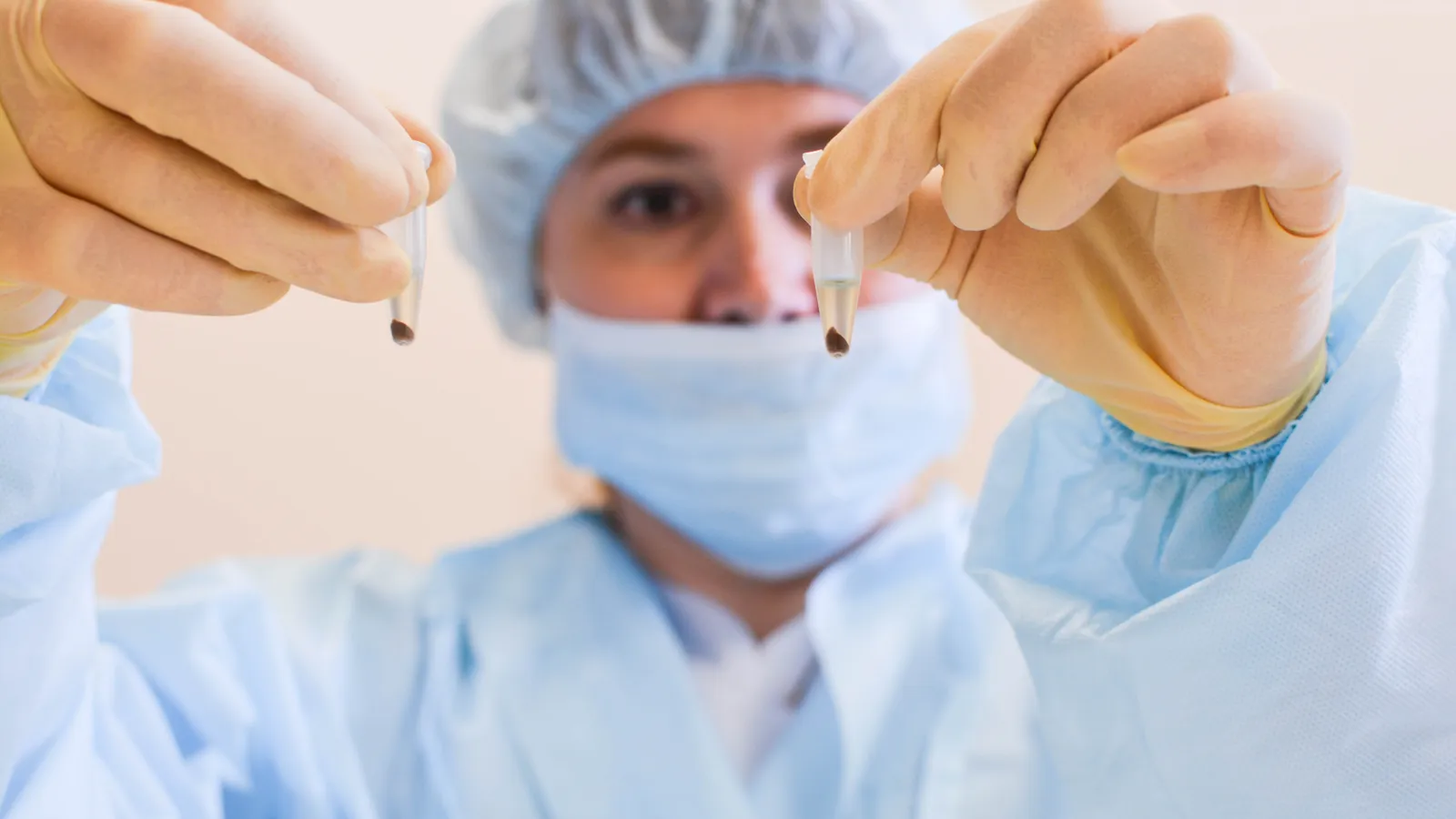 Lab worker in mask, cap and gown holds up two small sample tubes