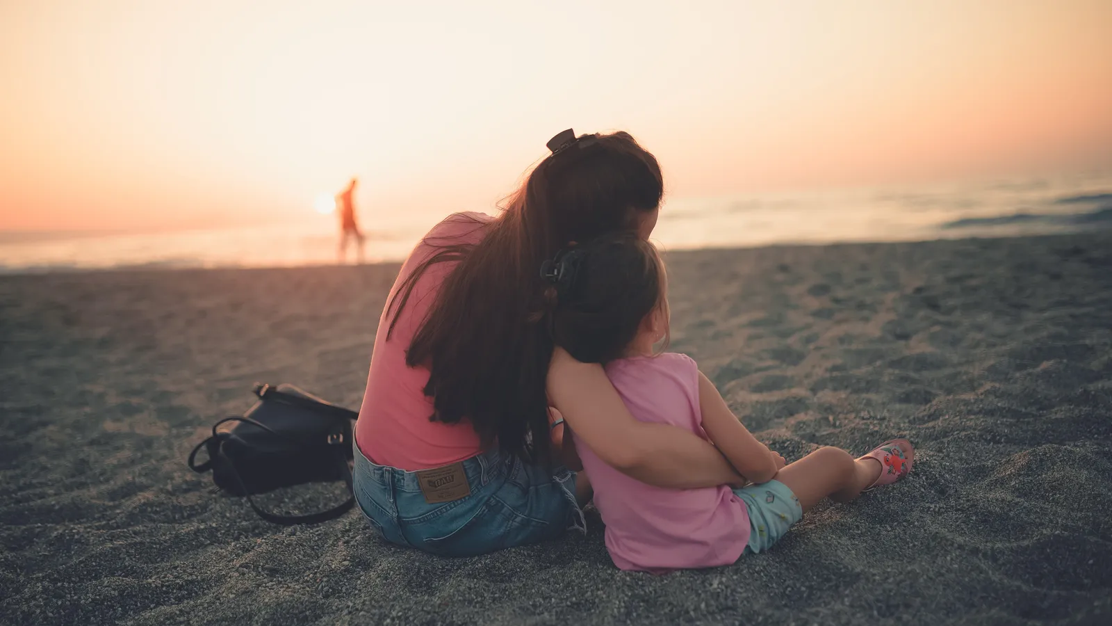 Mother and young daughter sitting on a beach at sunset, hugging, seen from behind