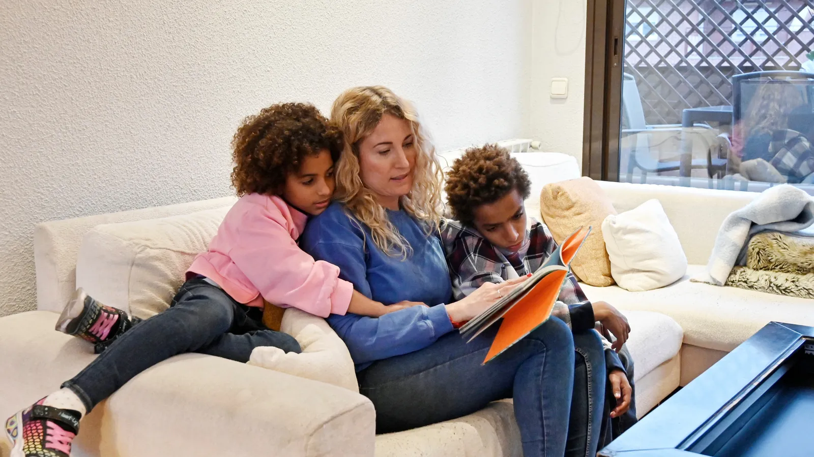 Mother on a sofa reading a book aloud with two children following along