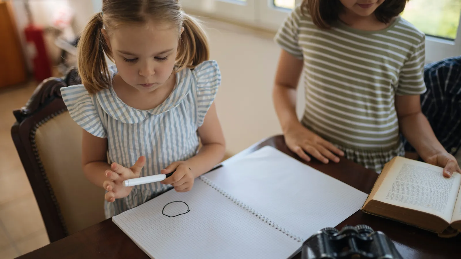 Older girl holding a book beside a younger girl drawing in a grid notebook