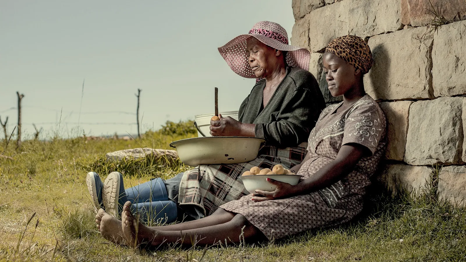 Older woman and young woman sitting side by side on grass against a stone wall, preparing peaches