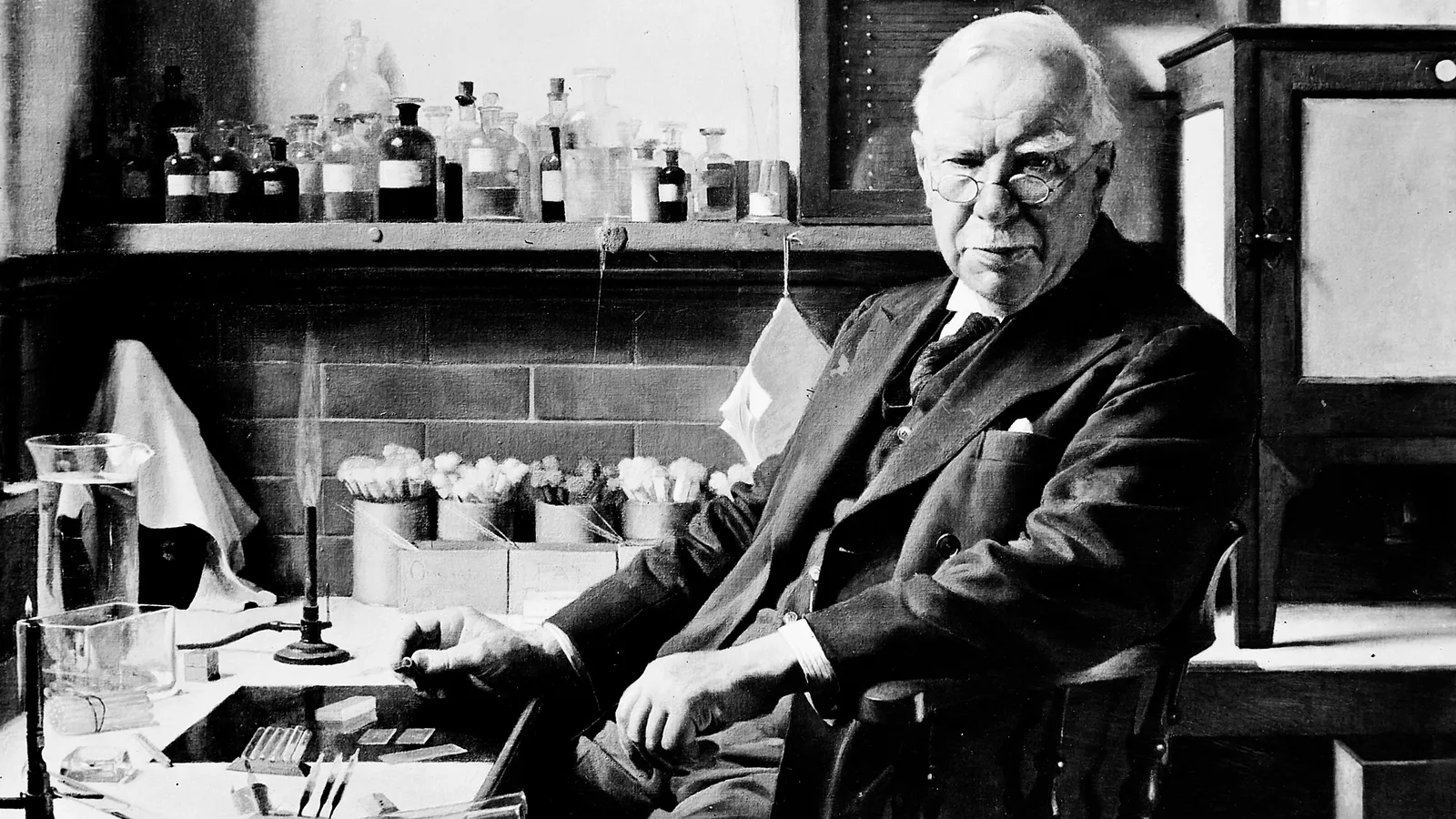 Painted portrait of an elderly Almroth Wright seated at a laboratory bench beside shelves of bottles