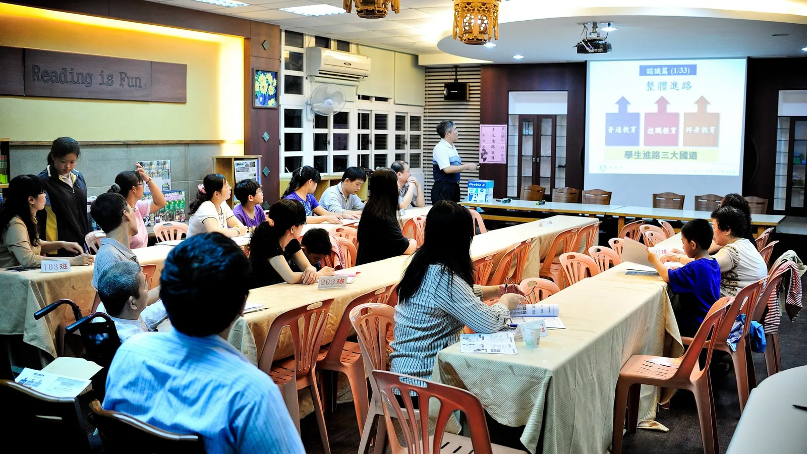 Parents and students seated at long tables while a teacher presents slides at a school meeting