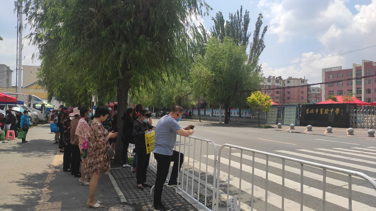 Parents standing at a metal barrier outside a school exam site, waiting