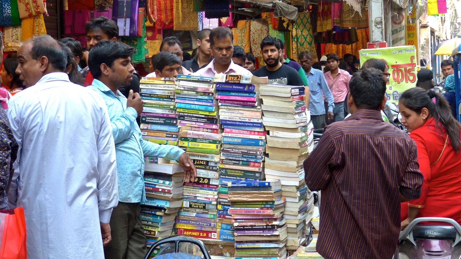 People around tall stacks of second-hand books at a busy Delhi street stall