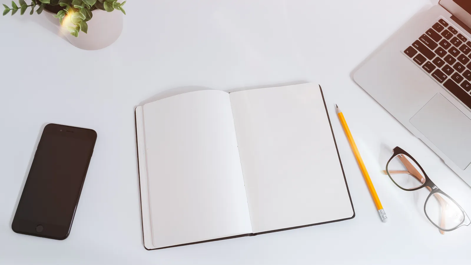 Phone with a dark screen beside an open blank notebook, pencil, glasses and laptop on a white desk