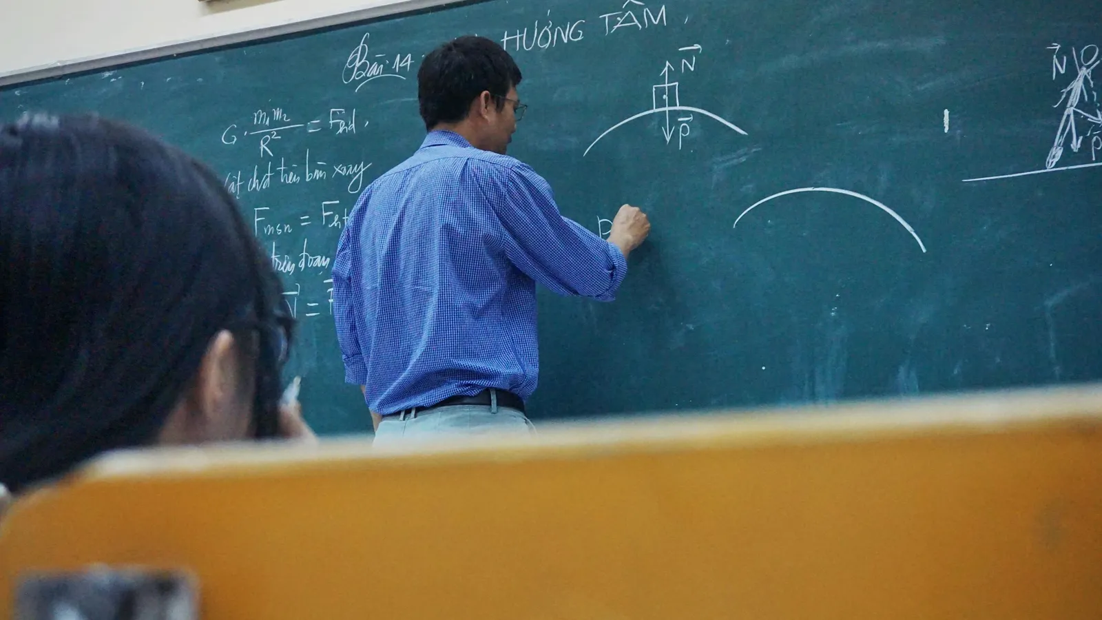 Physics teacher writing equations on a green chalkboard, seen from a student desk