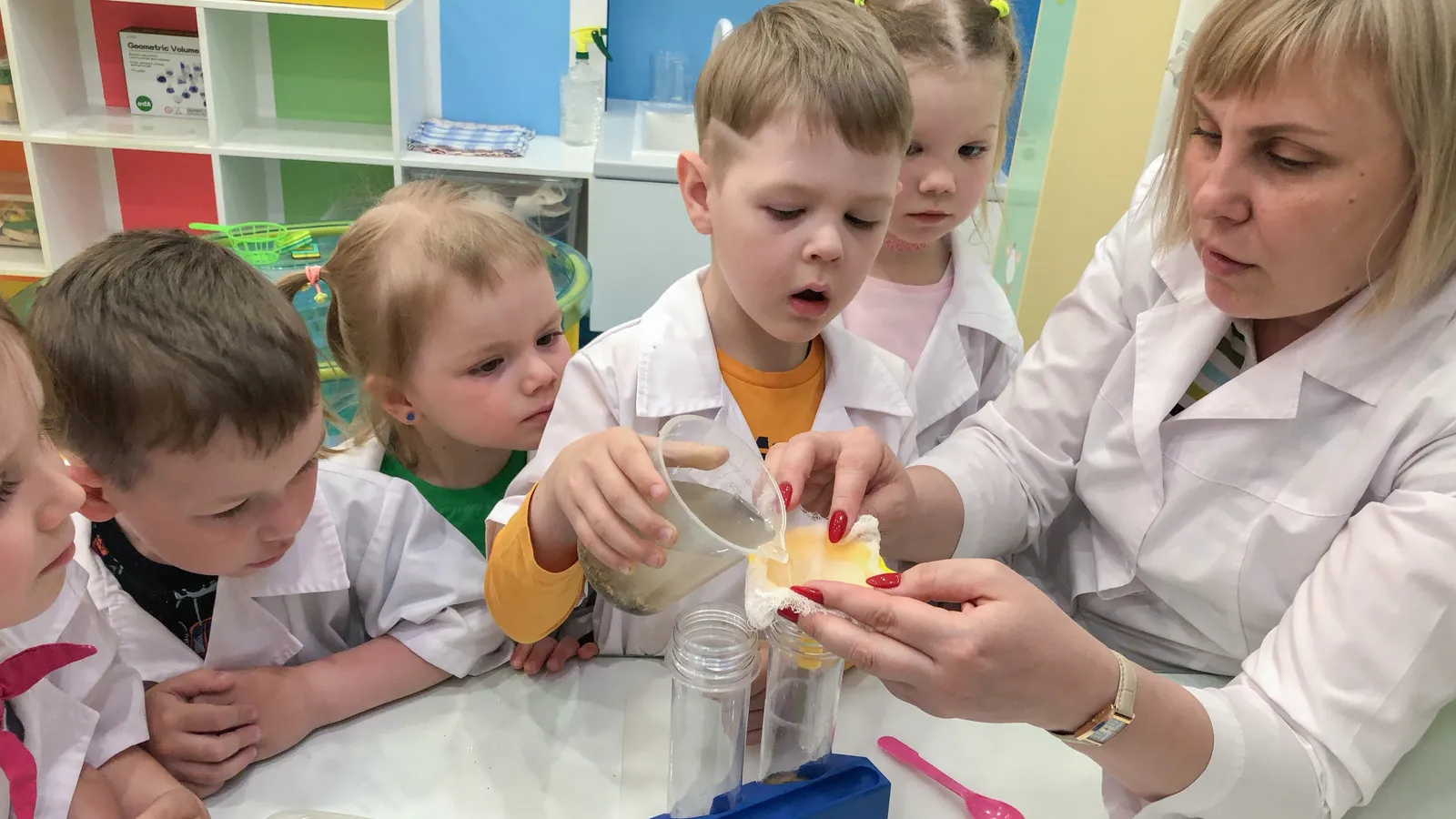 Preschoolers in lab coats watch a boy pour cloudy water through a cloth filter held by a teacher
