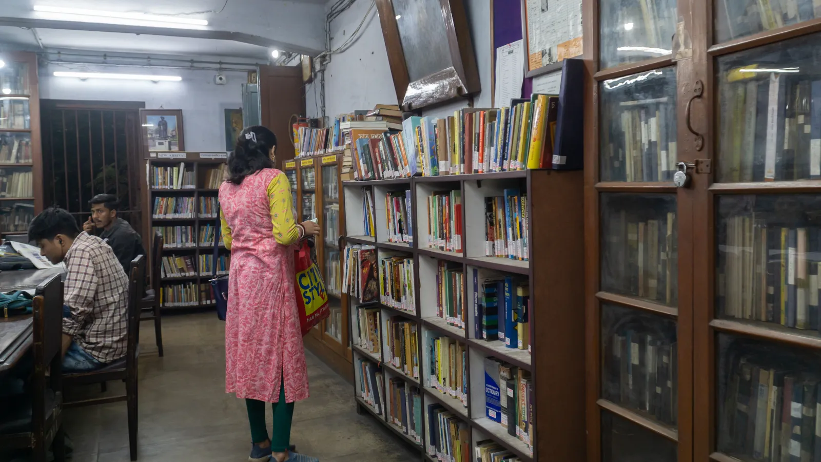 Readers at tables and a woman browsing bookshelves in a Kolkata public library