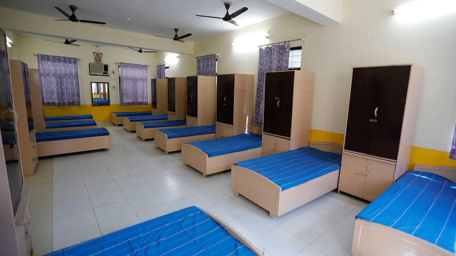 School hostel dormitory with rows of beds, blue sheets, wardrobes and ceiling fans