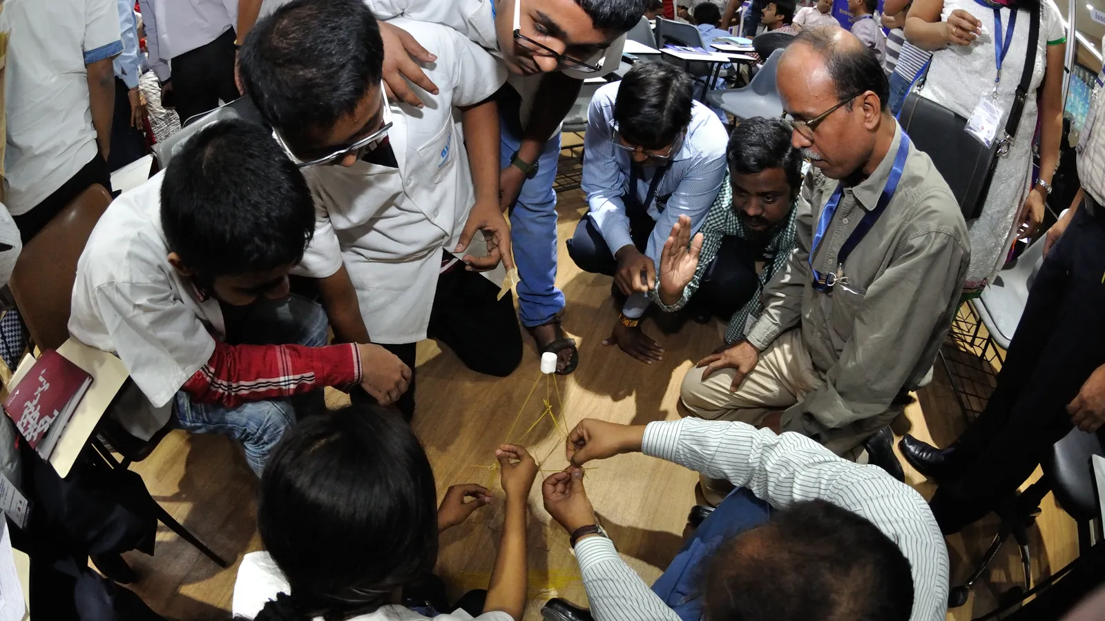 School students and adult workshop participants build a spaghetti and marshmallow tower on the floor