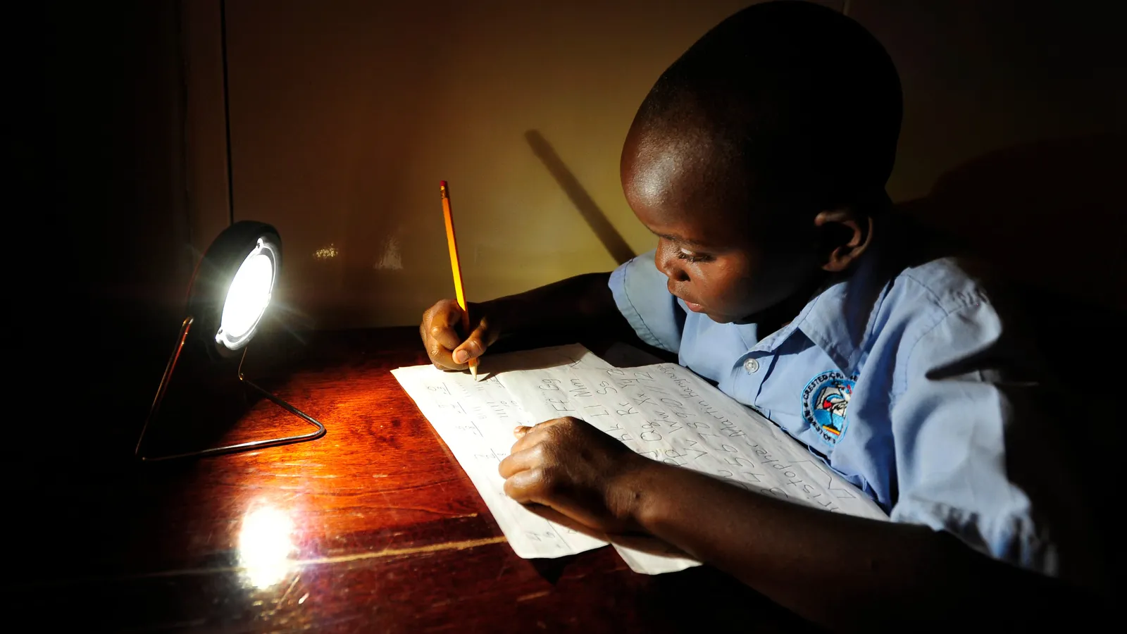 Schoolboy in a blue uniform writing with a pencil at a desk lit by a small solar lamp