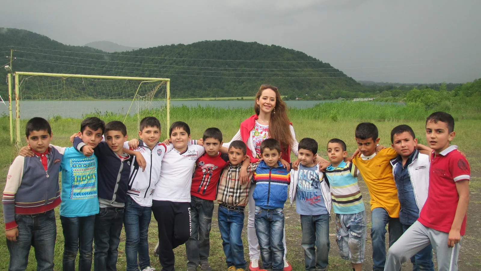 Schoolchildren and their teacher on a grassy football field by a lake and wooded hills