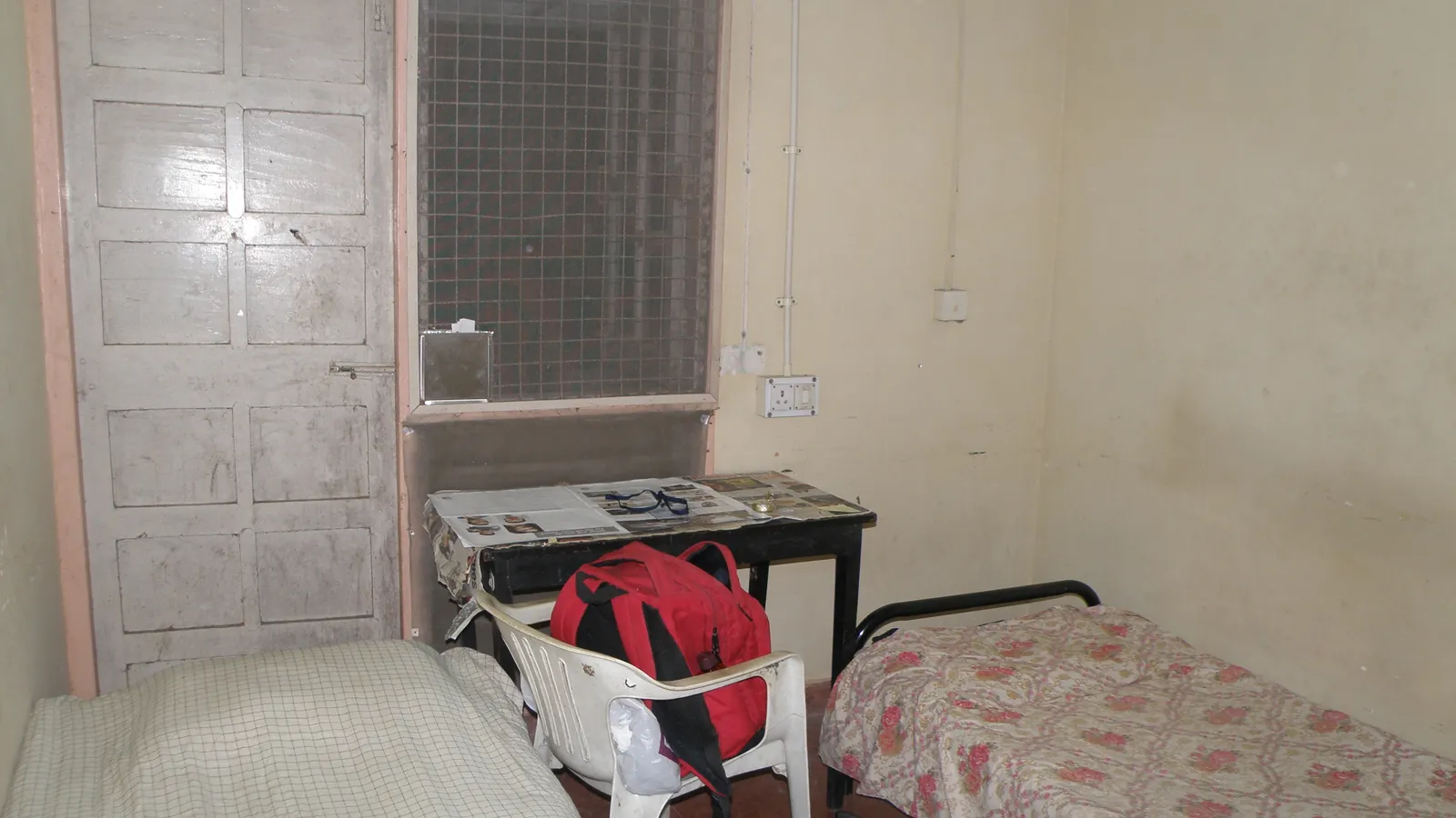 Shared Indian hostel room with two beds, a desk, a plastic chair and a red backpack
