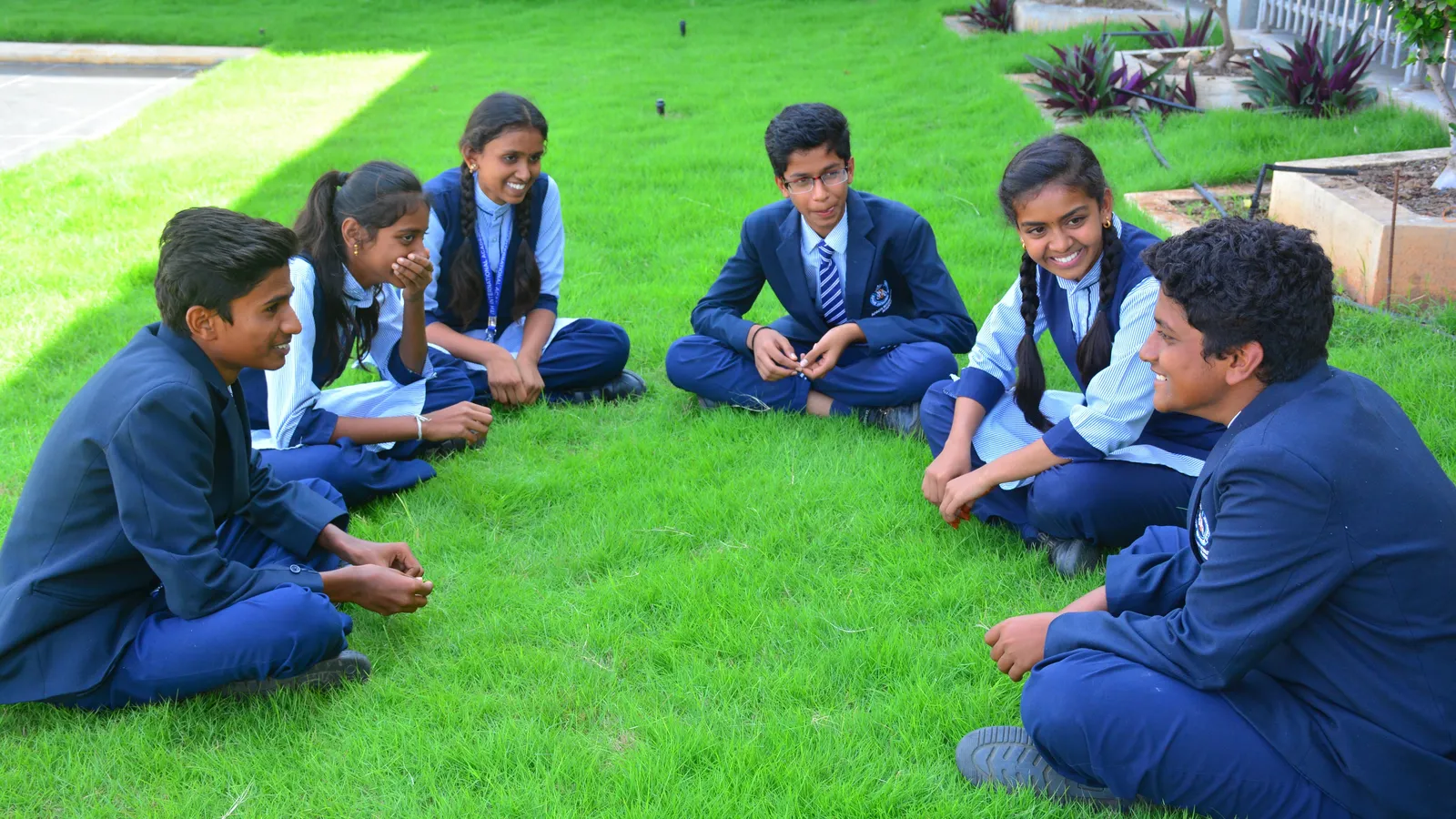 Six school students in navy uniforms sitting in a circle on grass, talking