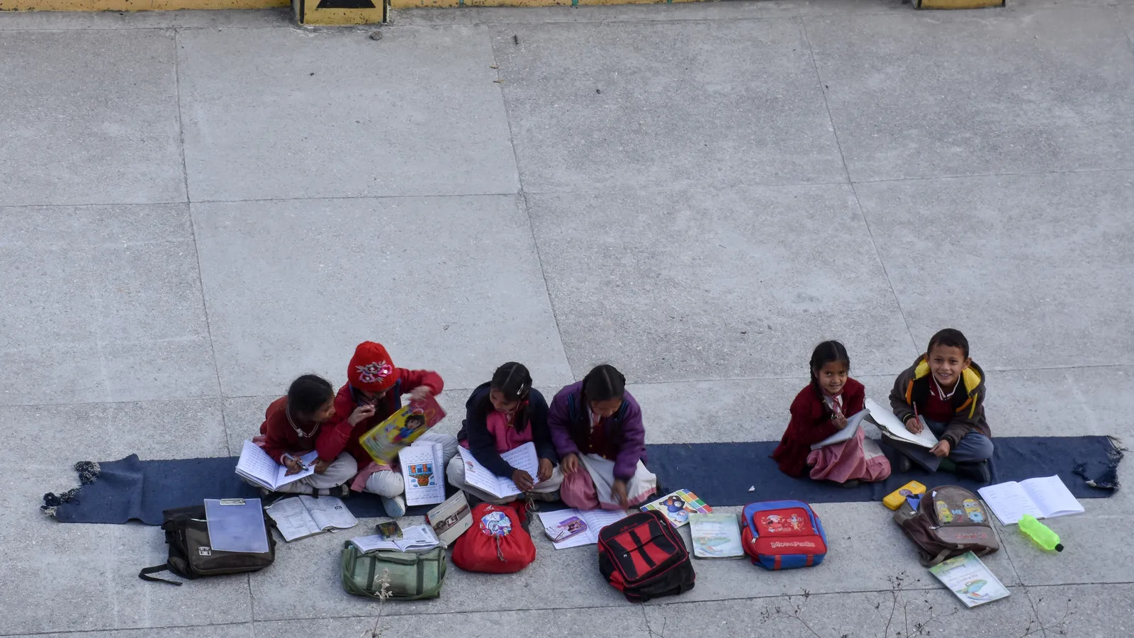 Six schoolchildren in uniform studying notebooks on a mat in a school courtyard
