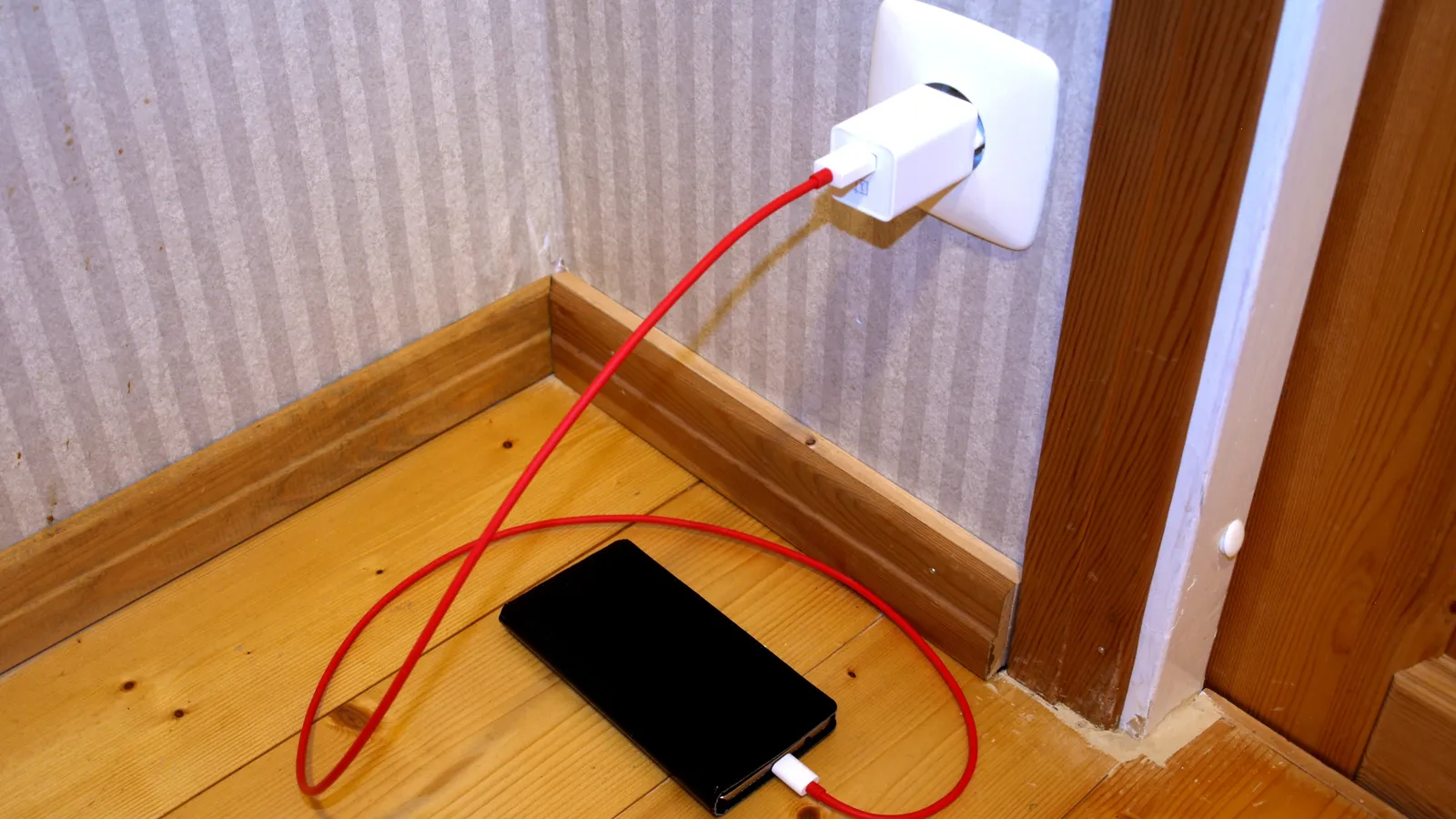 Smartphone charging on a wooden floor beside a doorway, red cable to wall socket
