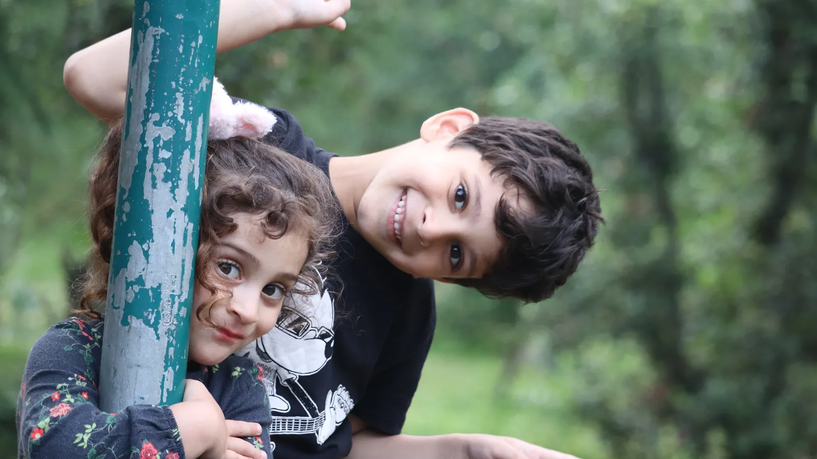Smiling brother and younger sister peeking around a green pole outdoors