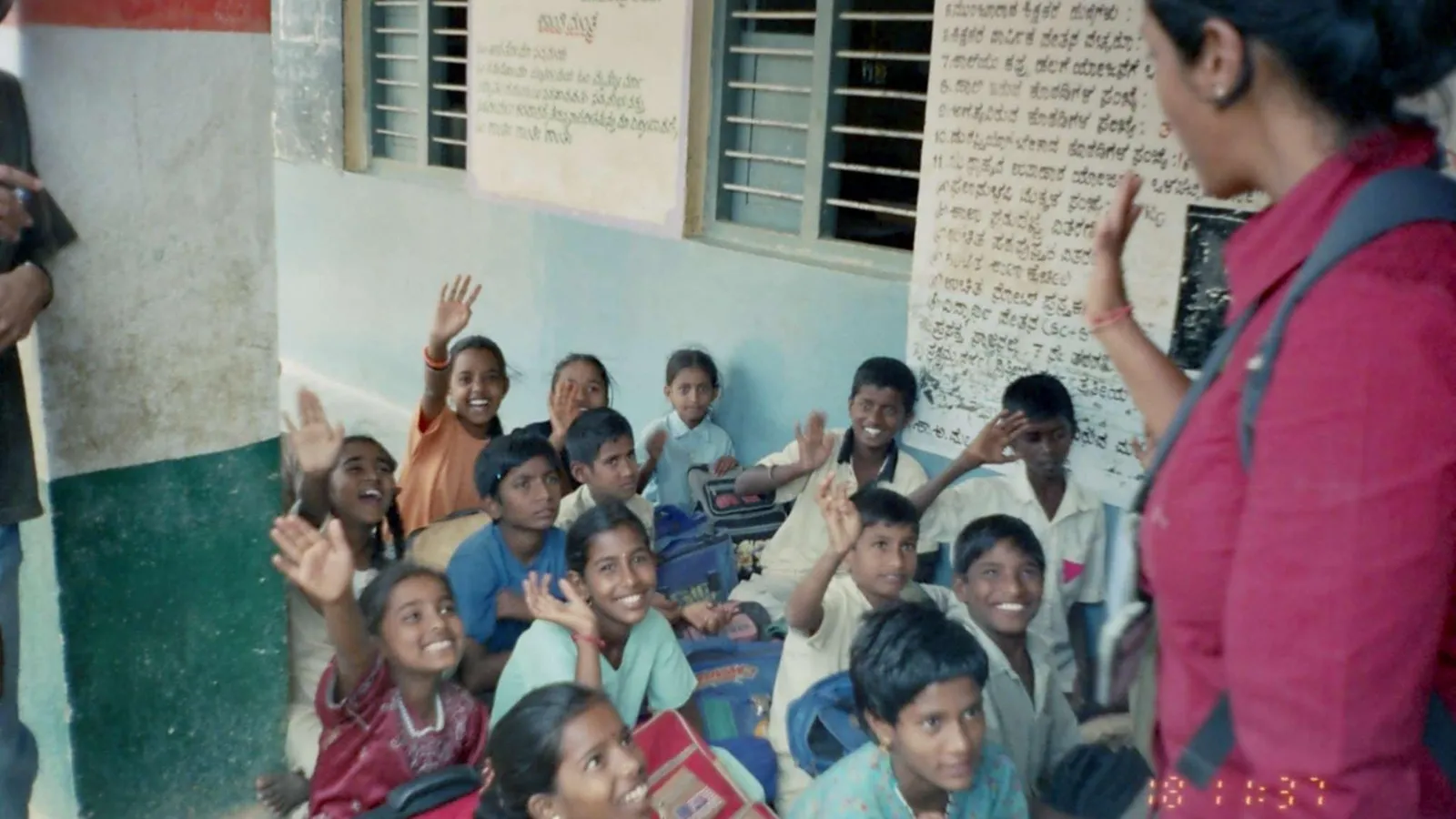 Smiling children seated on a school floor in Bangalore raising hands towards a woman