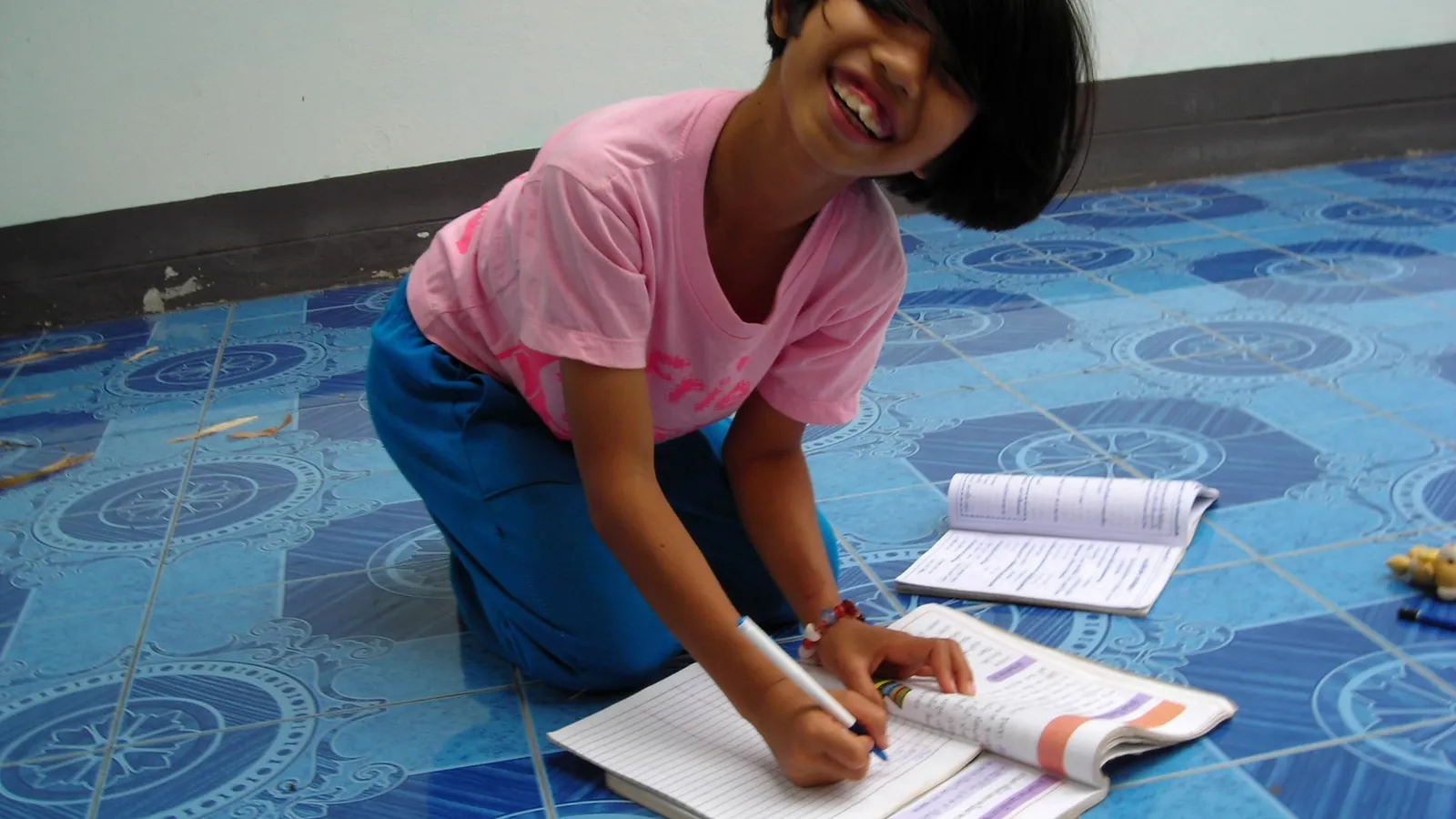 Smiling girl kneeling on a tiled floor, writing in her school notebook
