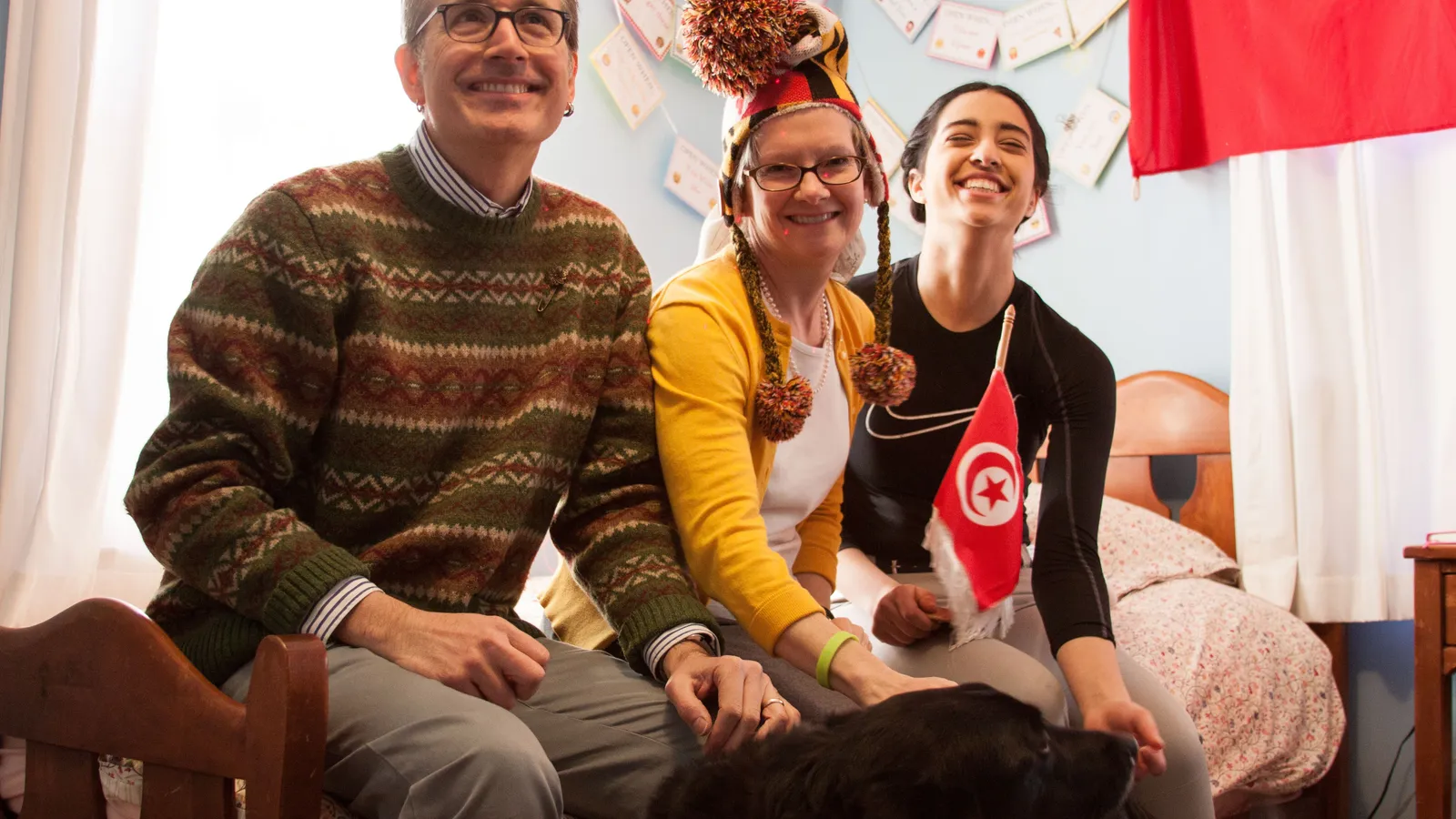 Smiling host parents and an exchange student holding a Tunisian flag, sitting on a bed with their dog
