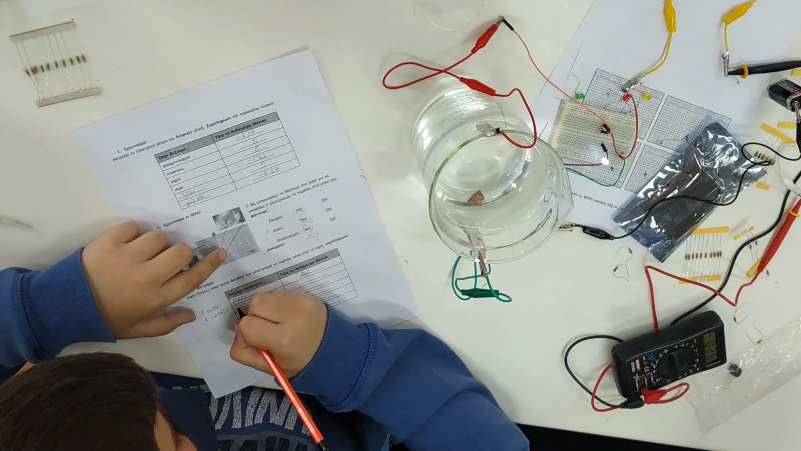 Student fills in a worksheet beside circuit boards, wires, a water beaker and a multimeter