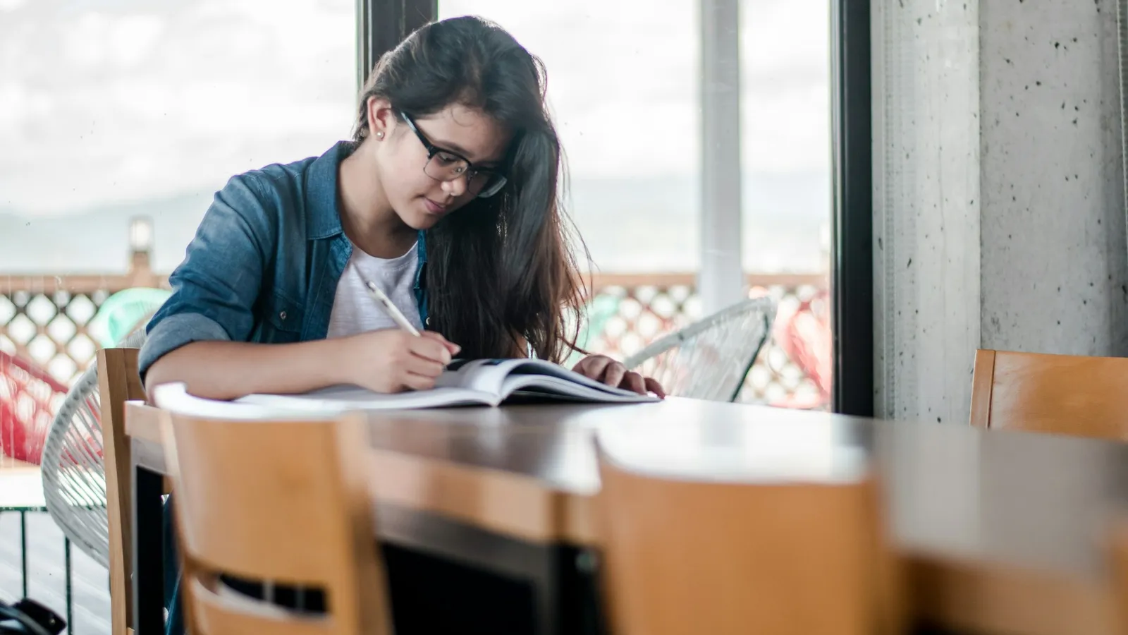 Student in glasses writing notes at a desk beside a bright window
