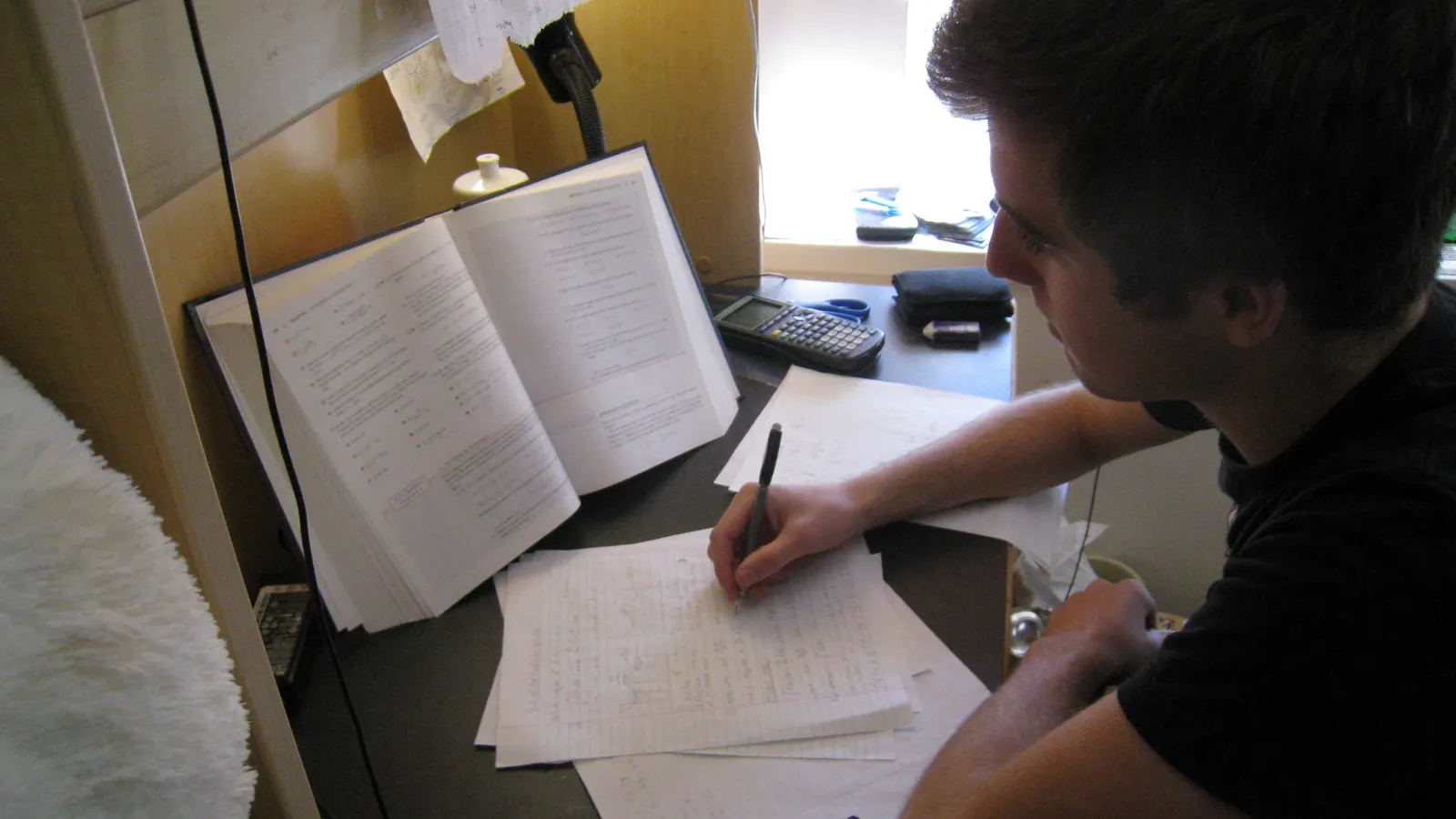 Student writing notes at a desk beside an open textbook and calculator