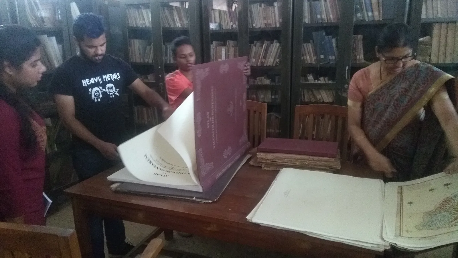 Students and a woman in a sari look through large old atlases in a university library