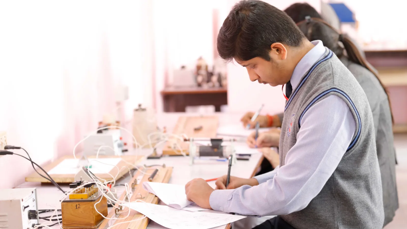 Students in school uniform writing observations at a physics lab bench with circuit apparatus