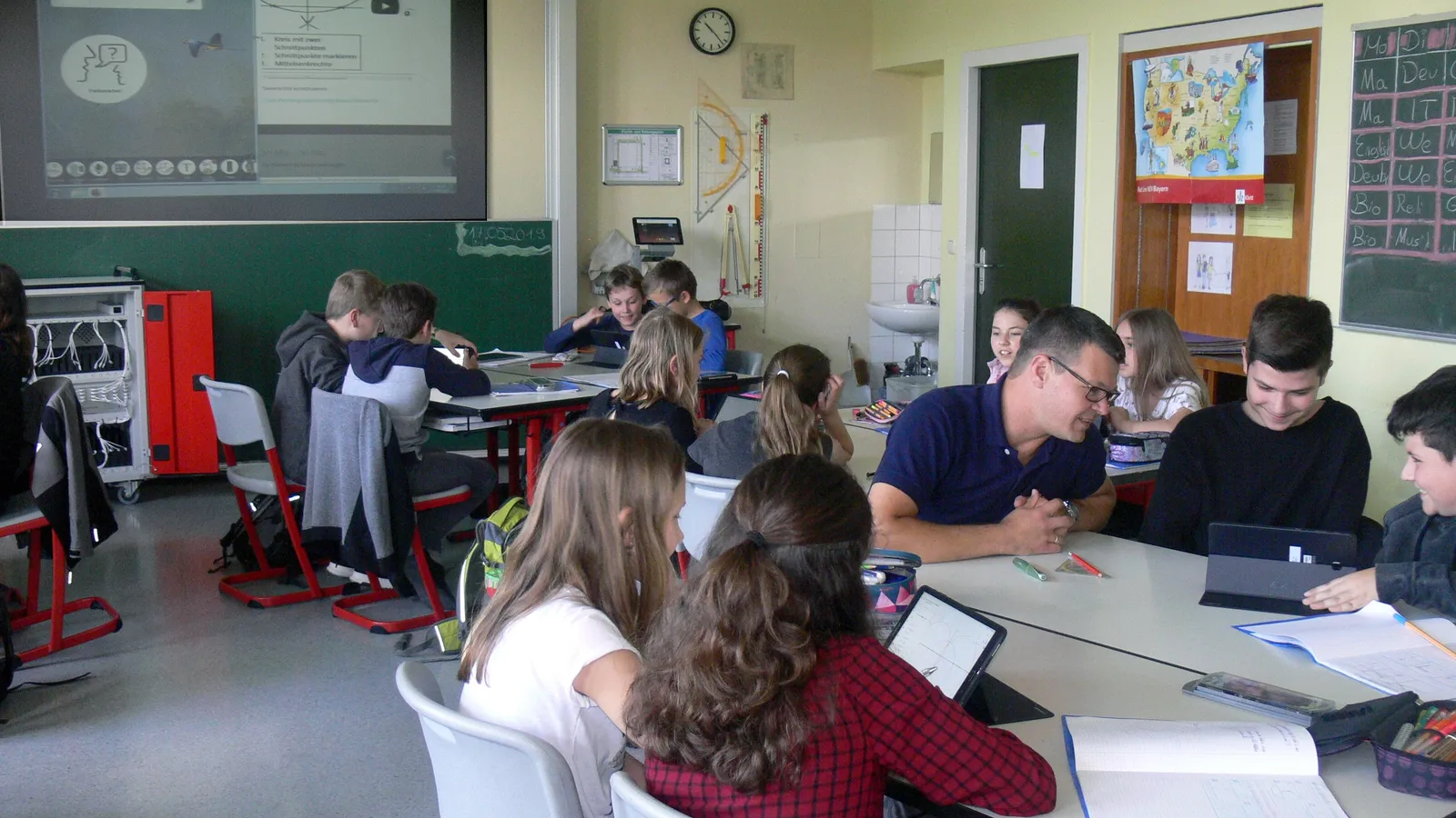 Students working on tablets at classroom tables while a teacher talks with them