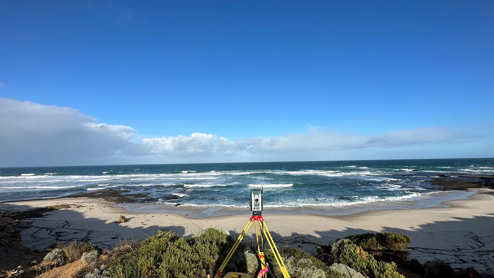 Surveying total station on a yellow tripod overlooking a beach and ocean