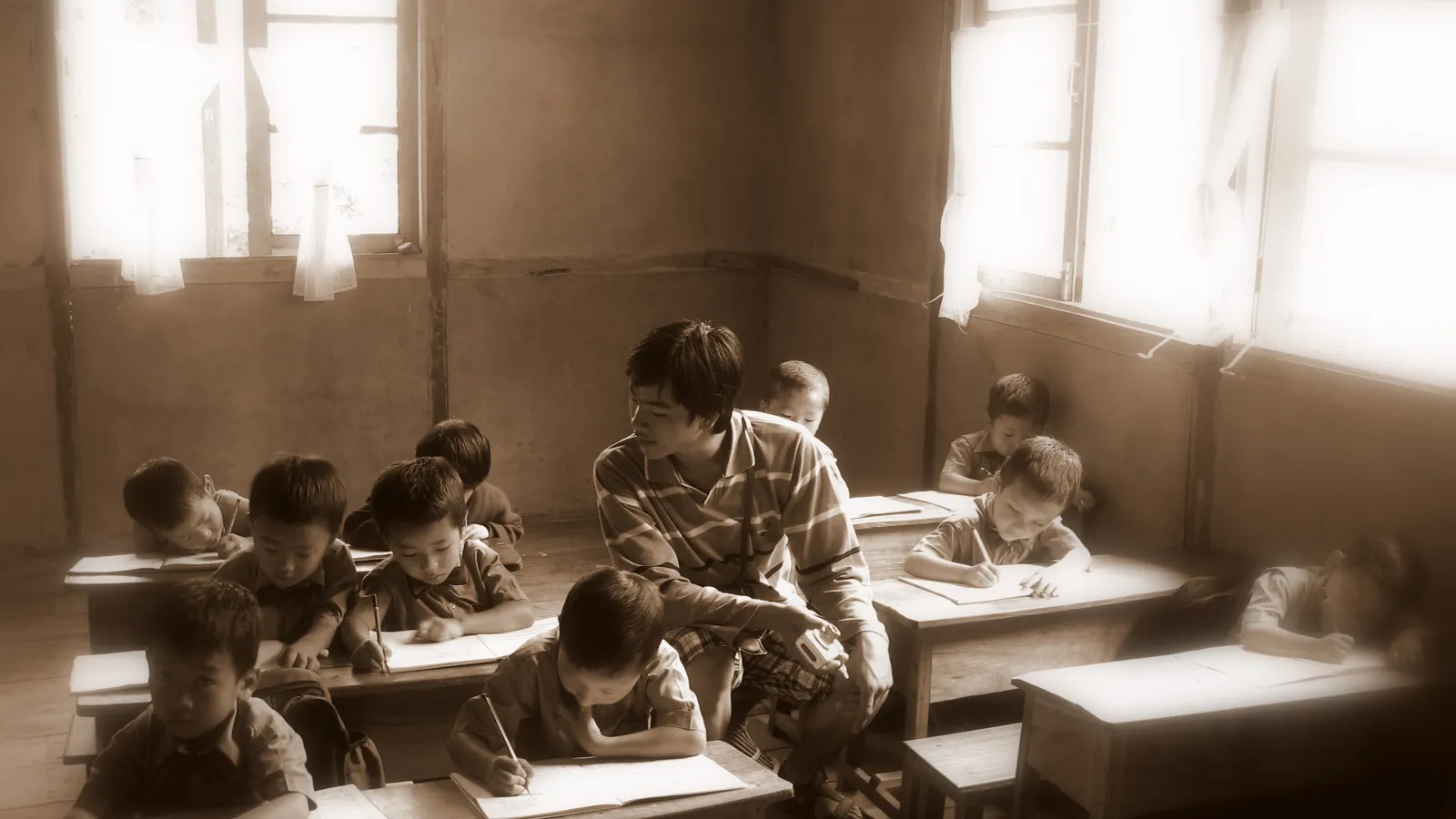 Teacher sits among young pupils writing in notebooks at wooden classroom desks