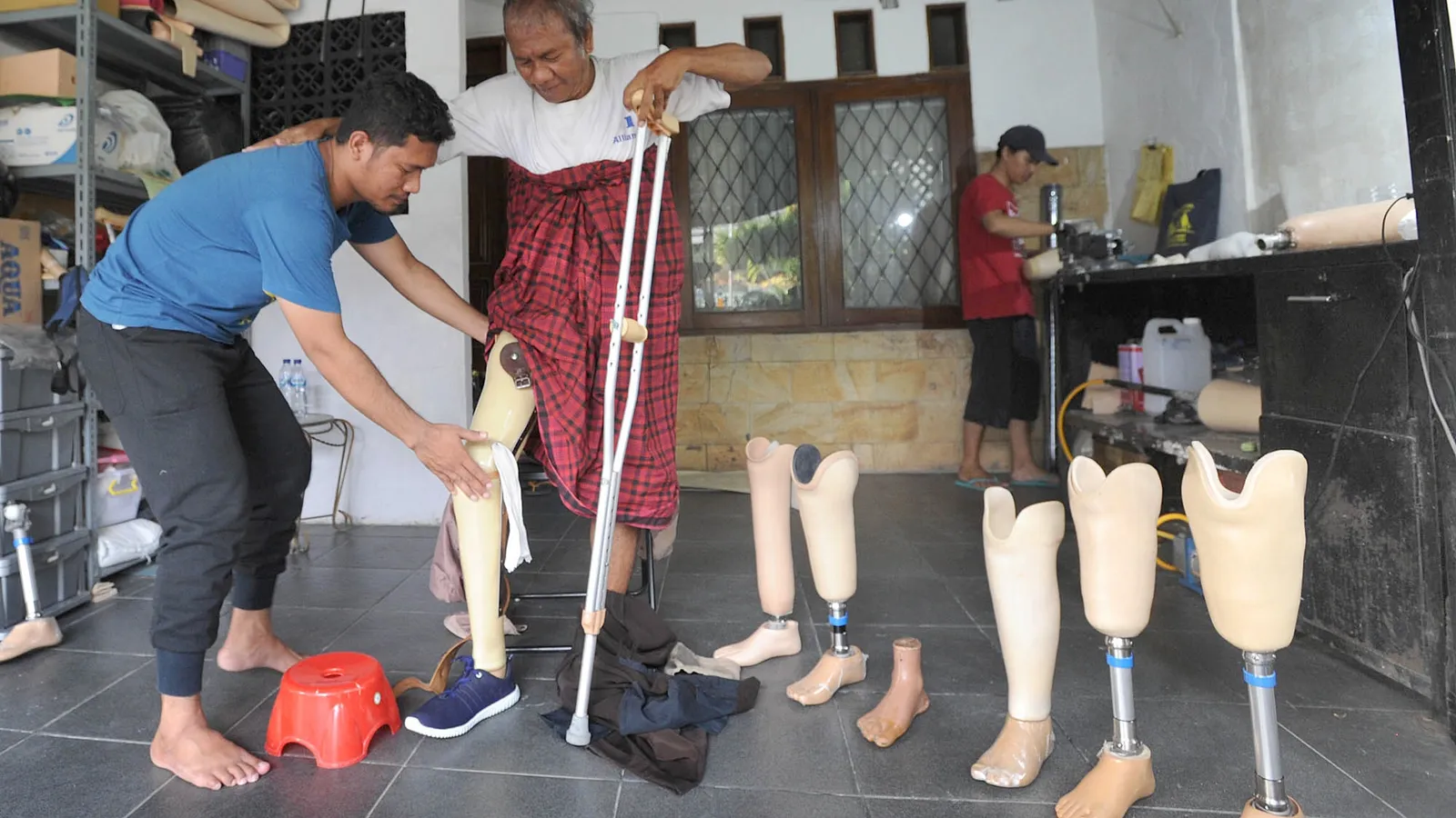 Technician adjusting an artificial leg on a man with crutches in a limb workshop