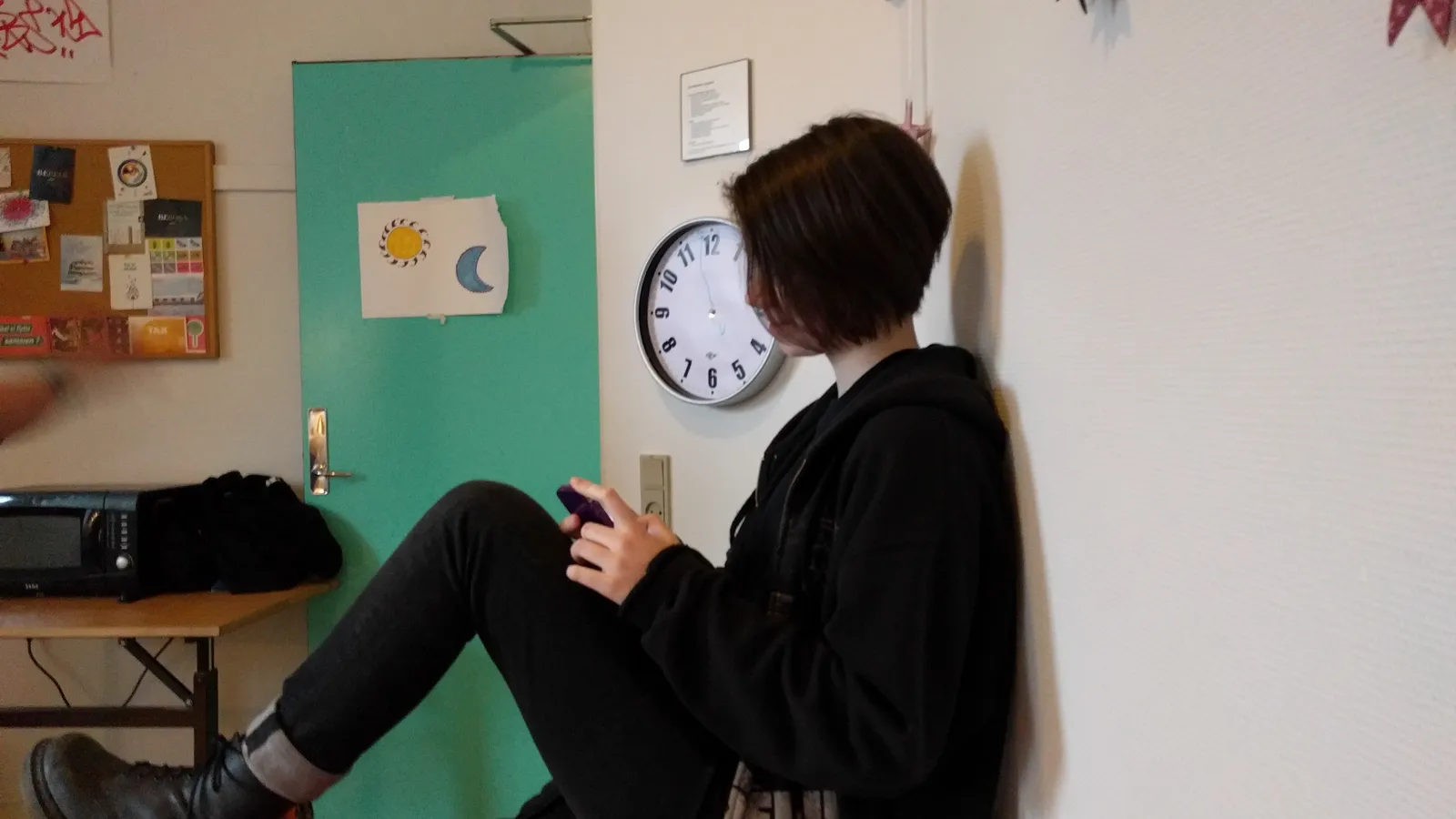 Teenager in a black hoodie sitting against a wall, looking at a phone