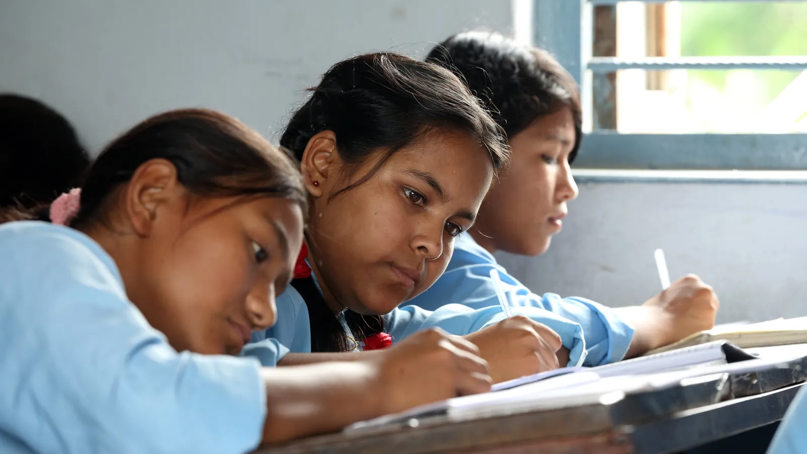 Three school students in blue uniforms writing in notebooks at a classroom desk