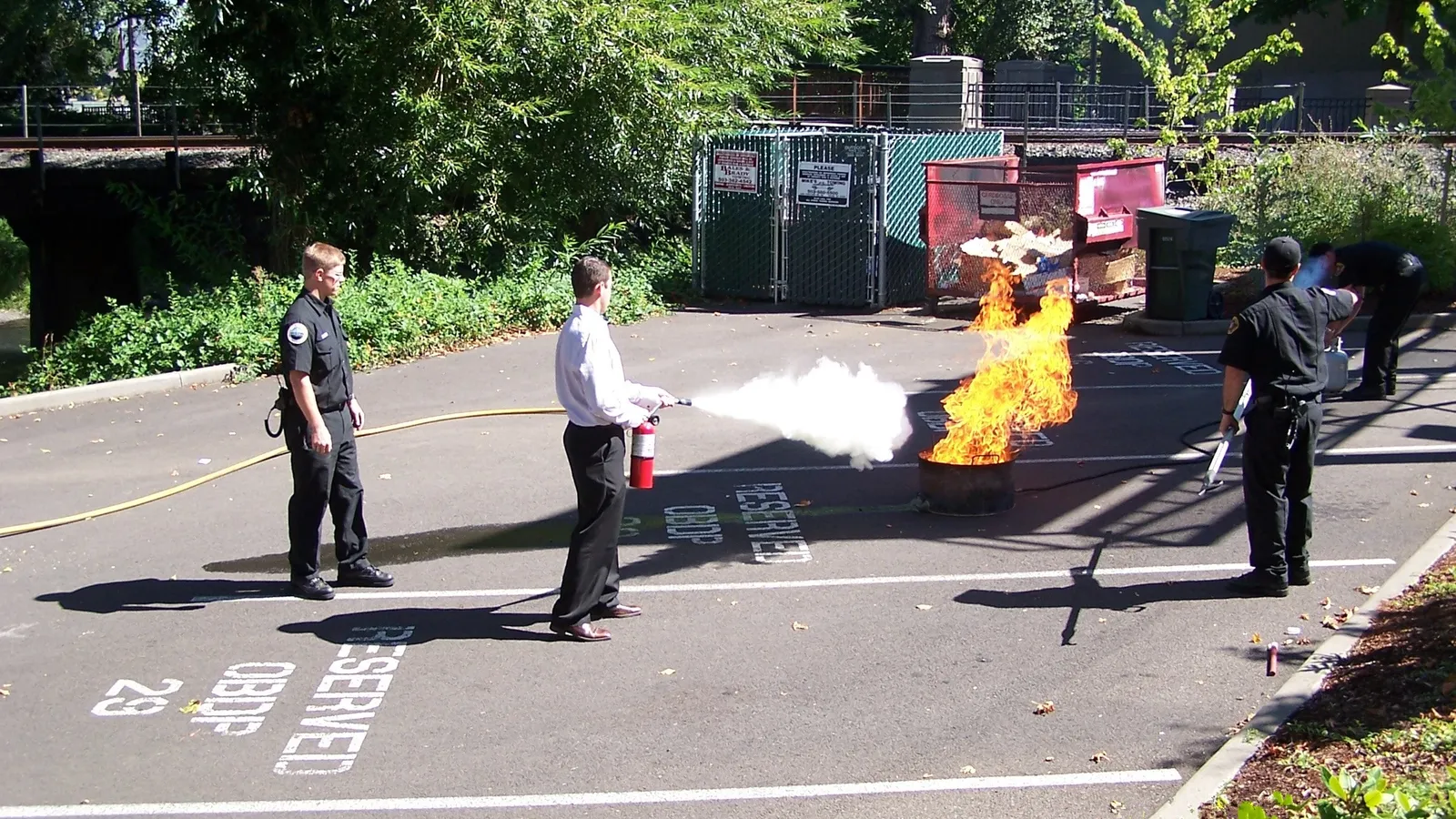 Trainee sprays a fire extinguisher at a fire in a metal tub while firefighters watch
