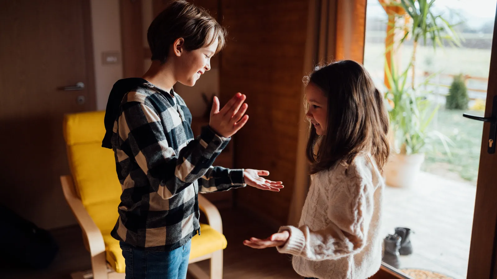 Two children play a clapping game indoors beside a glass door
