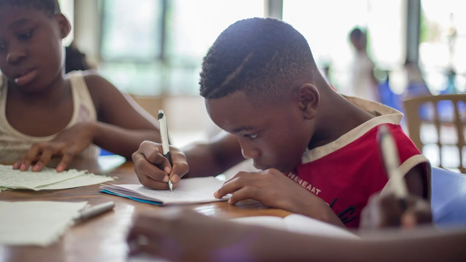 Two children writing in notebooks at a classroom table