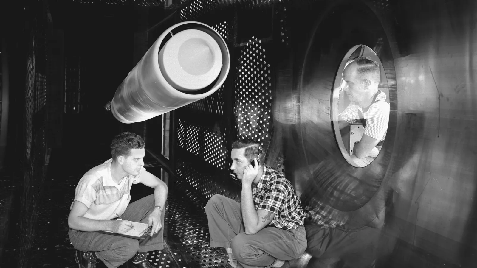 Two engineers crouch beneath a model in a wind tunnel as another watches