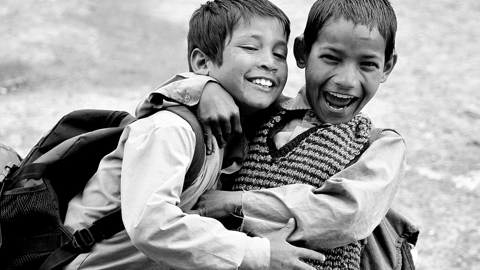 Two laughing schoolboys with backpacks hugging each other, black and white