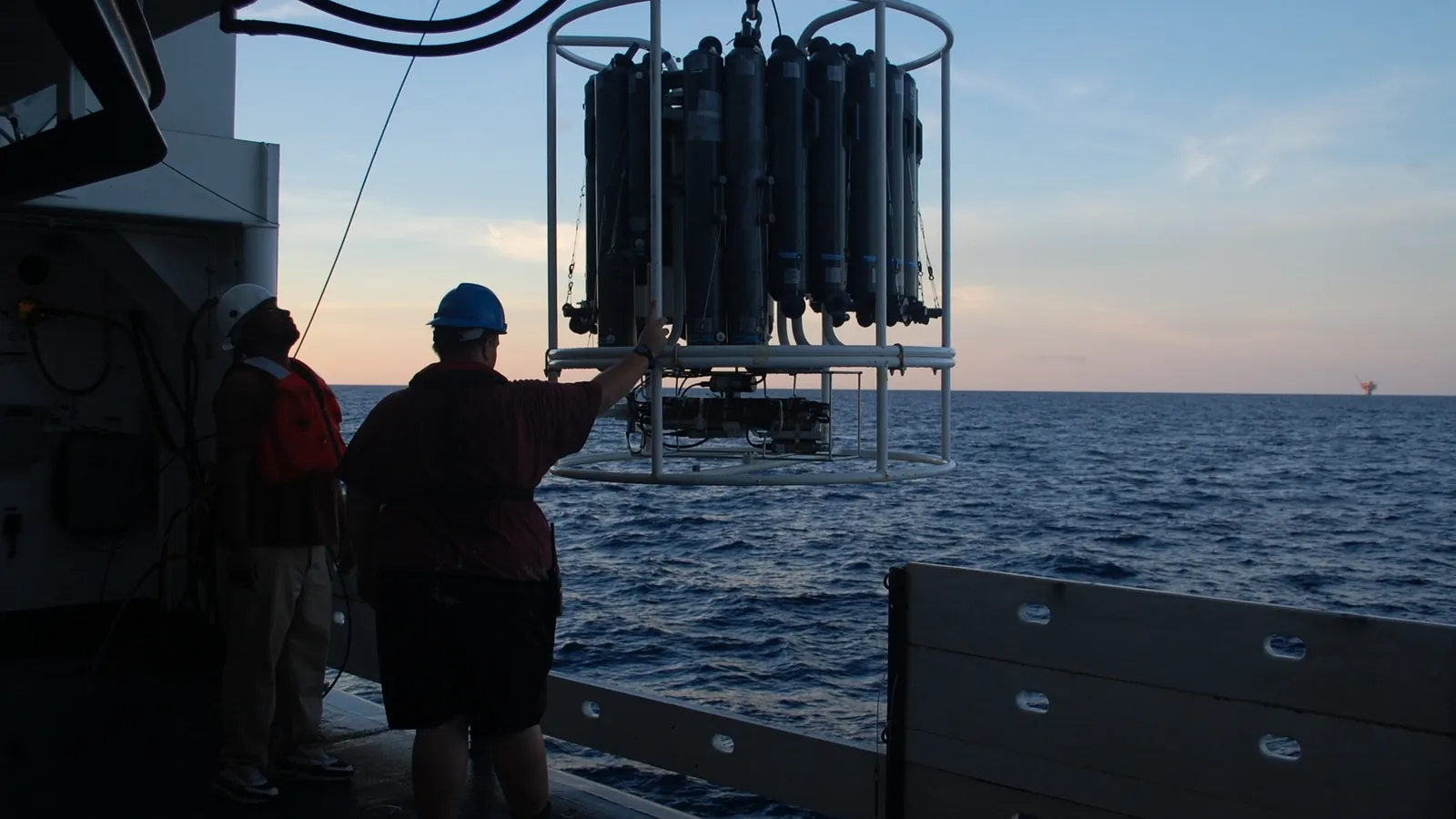 Two researchers in hard hats guide a CTD water sampler off a ship's deck at sunset