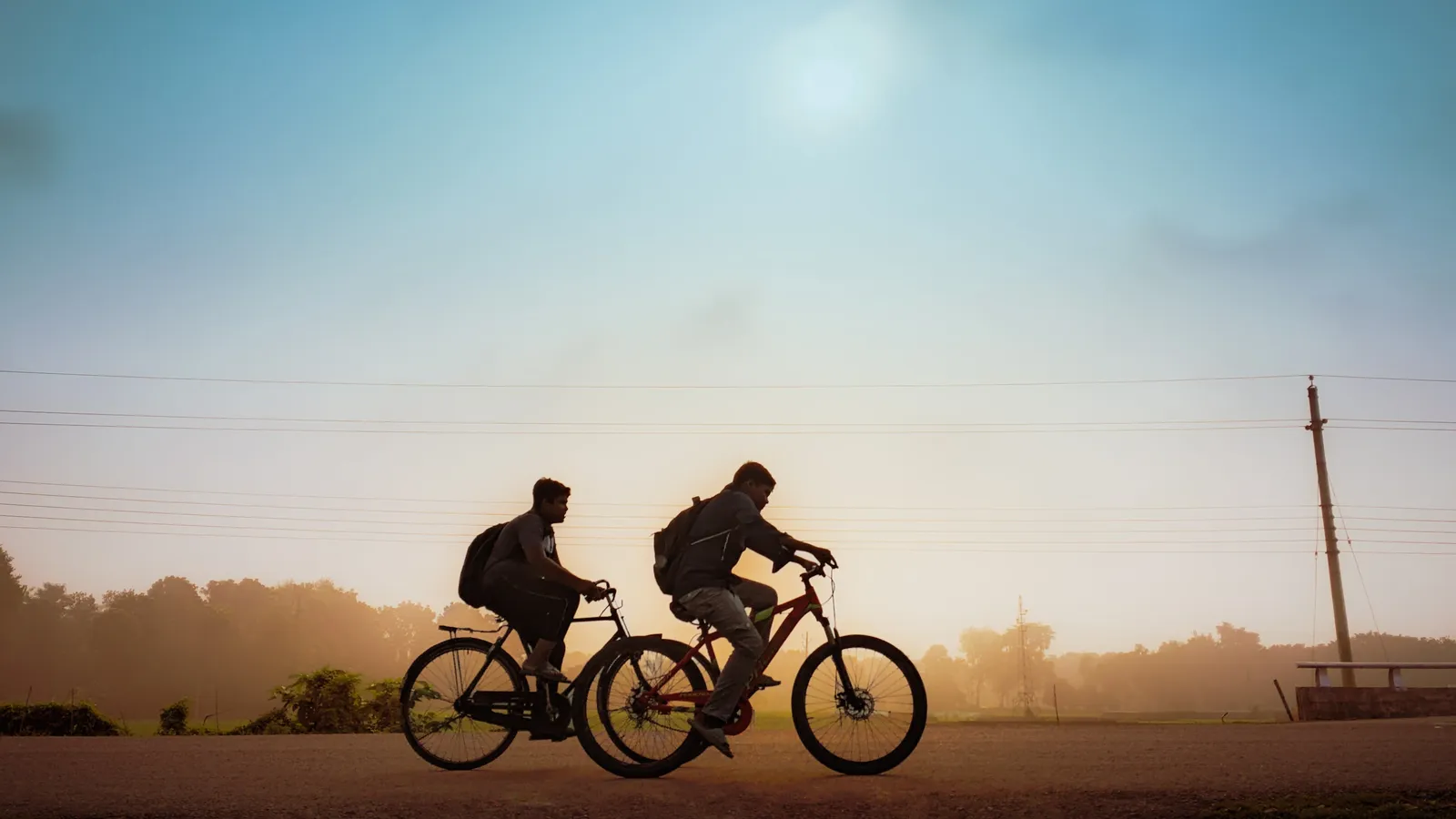Two students with backpacks cycling along a rural road in morning mist